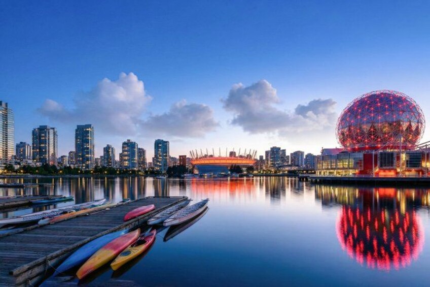 A waterfront cityscape with a large spherical building reflecting on calm water, docked kayaks, and modern high-rise buildings. 4