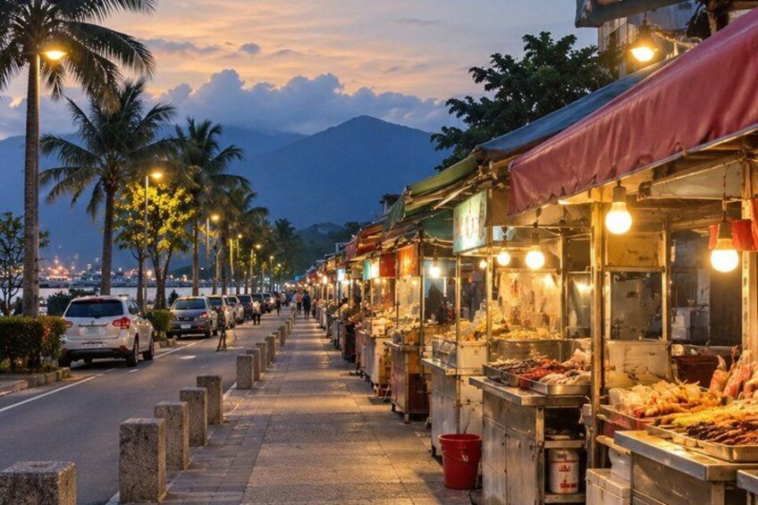 A street lined with food stalls at dusk, palm trees, and a mountain in the background. 4