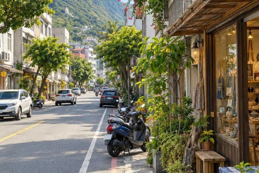 A street lined with parked cars and motorcycles, a row of shops with visible merchandise, and a mountain in the background. 2