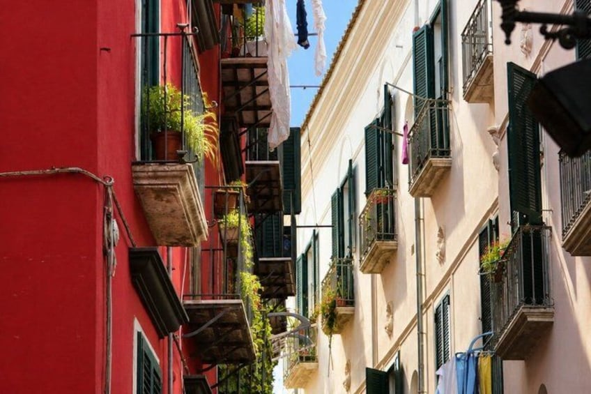A narrow street with buildings featuring balconies with plants and laundry hanging. 3