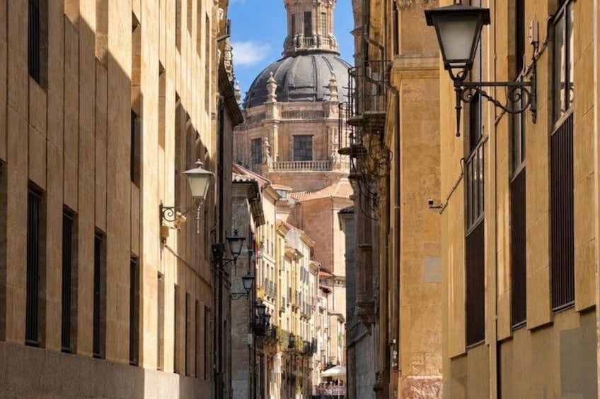 A narrow street with buildings on either side, a large dome structure in the background, and street lamps lining the path. 1