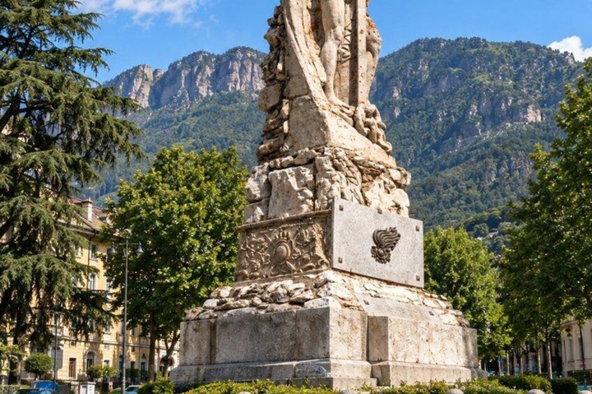 A stone monument with a lion's head, surrounded by trees and buildings, with mountains in the background. 3