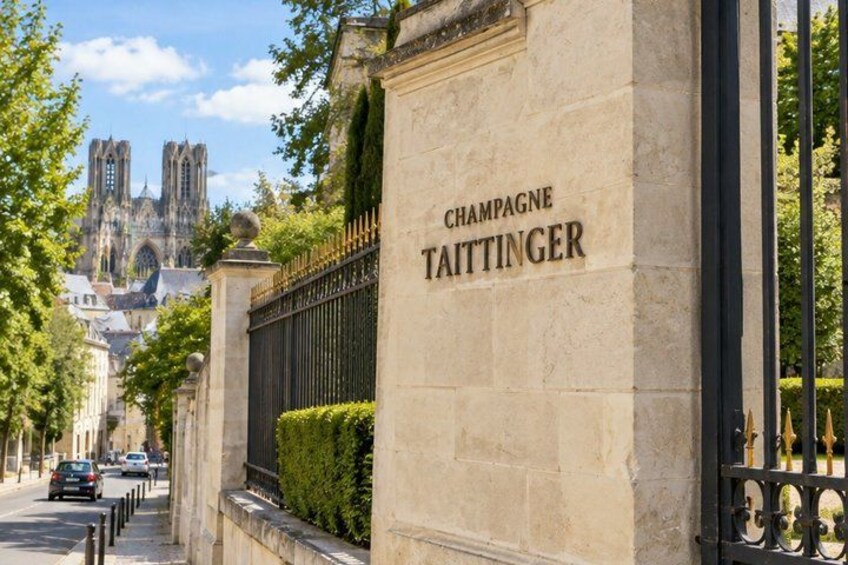 A stone wall with 'Champagne Taittinger' engraved on it, a wrought iron gate, and a cathedral in the background. 3