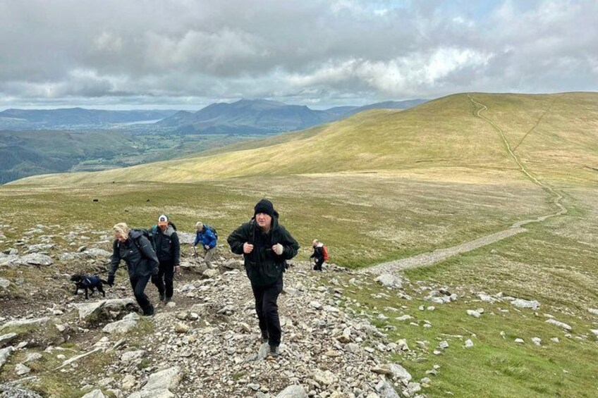 A group of hikers on a rocky mountain trail with a view of distant hills and a cloudy sky. 1