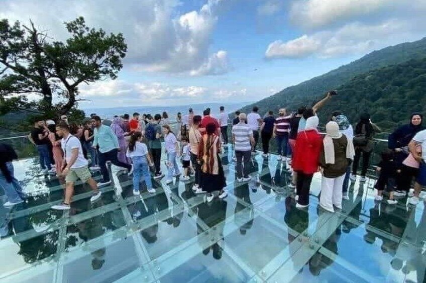 People standing on a glass bridge with a mountainous landscape in the background. 1