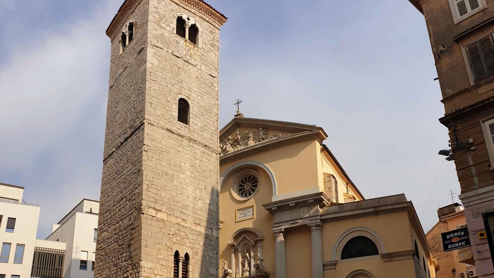 A historic stone tower with arched windows and a bell, adjacent to a building with a dome and a statue. 2