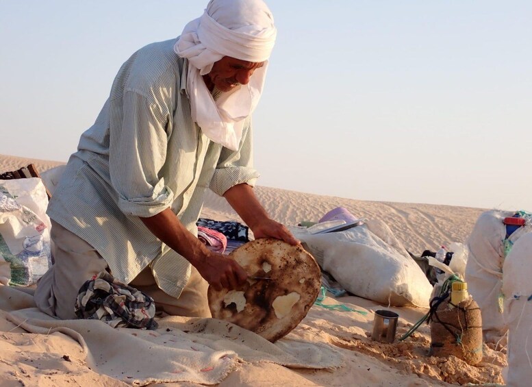 Une personne à genoux sur le sable, tenant un grand objet rond et plat, qui pourrait être un plat traditionnel, avec des sacs et un gobelet en métal à proximité. 5