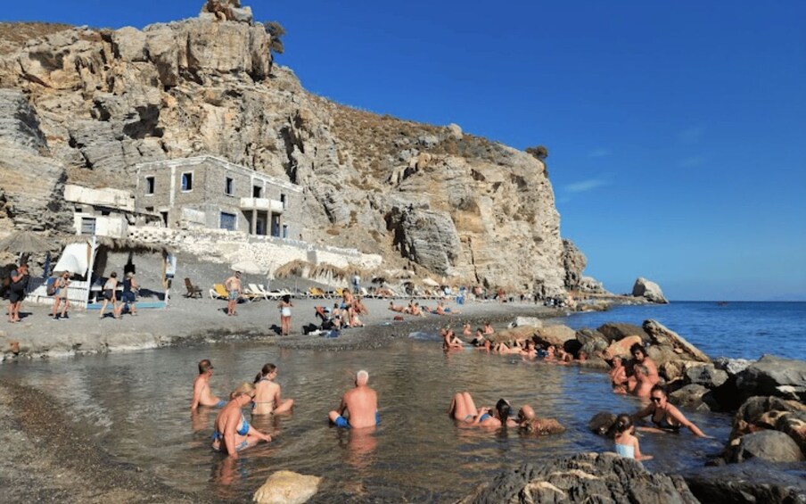 Une plage rocheuse où l’on voit des gens nager et bronzer, un bâtiment à la façade blanche et un ciel bleu dégagé. 2