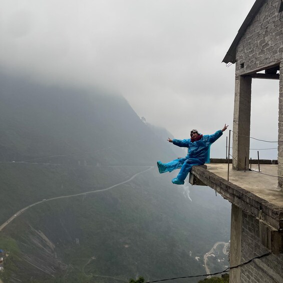 Une personne vêtue d’un imperméable bleu est assise sur une bordure en béton, les bras tendus, et contemple une vallée où serpente une route, avec une montagne en arrière-plan. 2