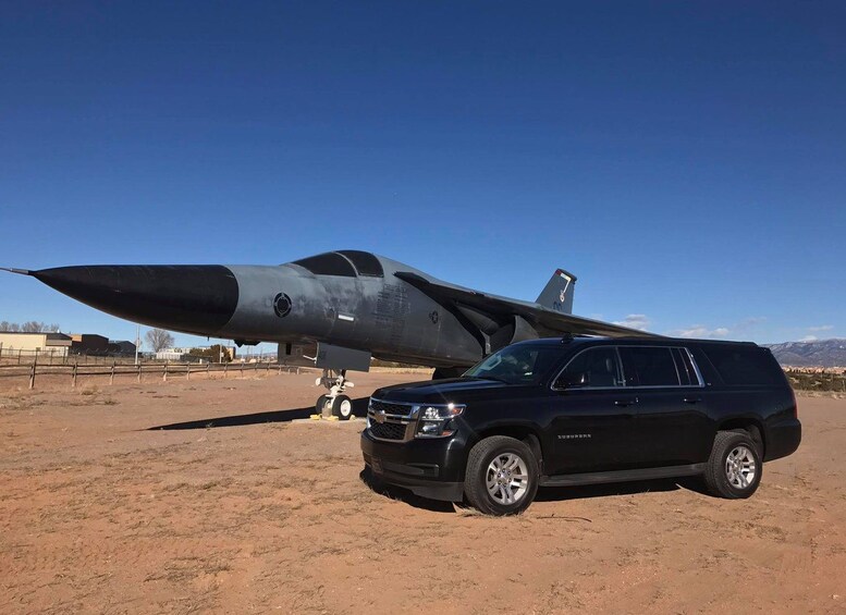 Un SUV negro estacionado en un desierto con un avión militar de fondo. 3