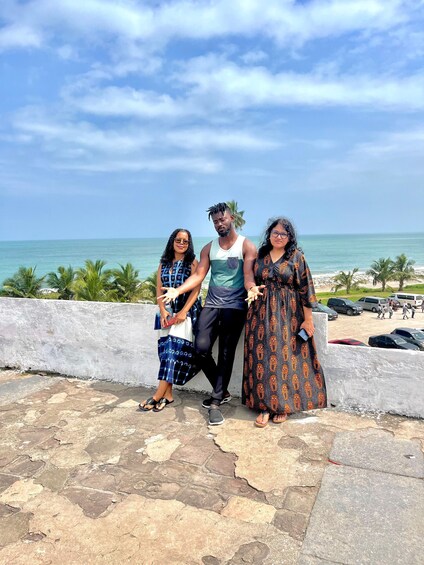 Three people standing on a stone platform with a beach and palm trees in the background. 3