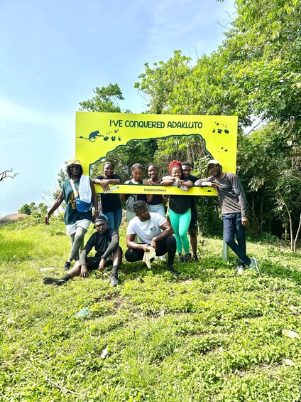 A group of people posing in front of a yellow sign with wildlife silhouettes and the text 'I'VE CONQUERED ADAKLUTO'. 1