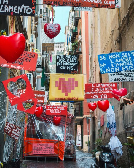 Una calle estrecha con varios carteles y decoraciones, incluyendo globos con forma de corazón y pancartas con mensajes. 3