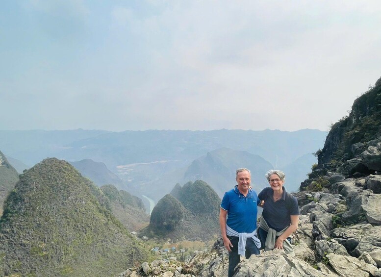Two people standing on a rocky mountain ridge with a valley and distant mountains in the background. 1