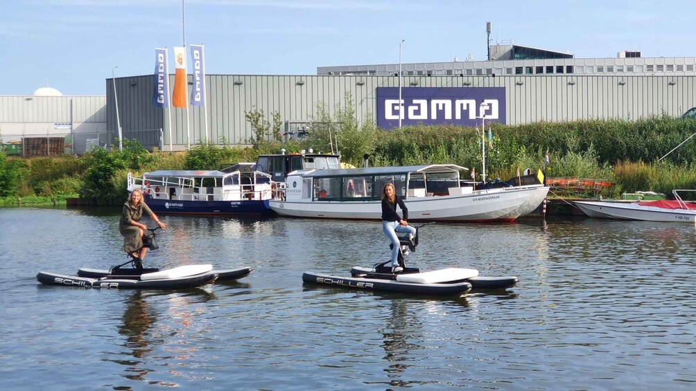 Två personer åker stående på en bräda på en lugn flod, med båtar och industriella byggnader i bakgrunden. 5