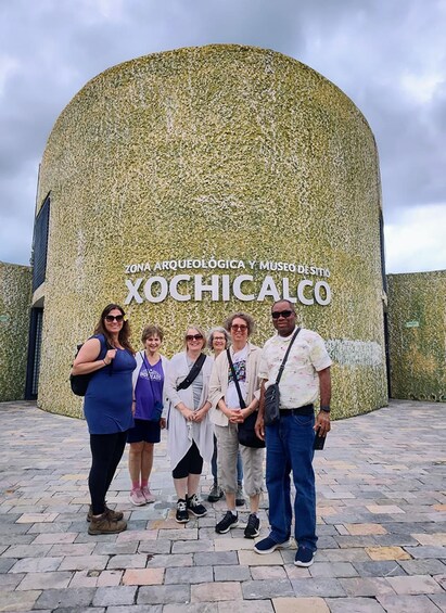 A group of people standing in front of a building with a textured facade and the text 'Zona Arqueológica y Museo de Xochicalco'. 3