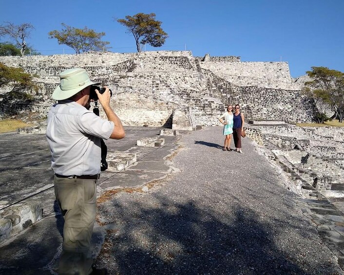 A person taking a photograph of a historical stone structure with two people standing in the foreground. 2