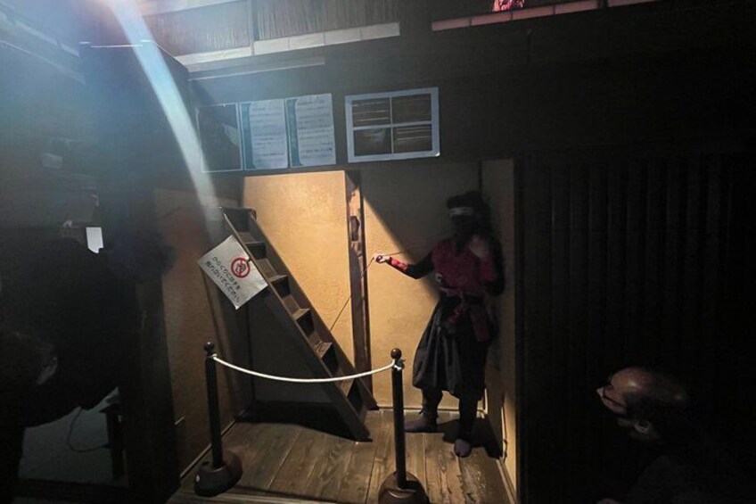 A person in a costume standing next to a staircase with a sign, in a dimly lit room with wooden walls. 4