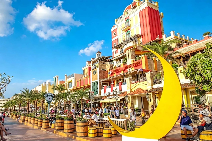 A bustling street with colorful buildings, palm trees, and a large yellow crescent moon sculpture. 4
