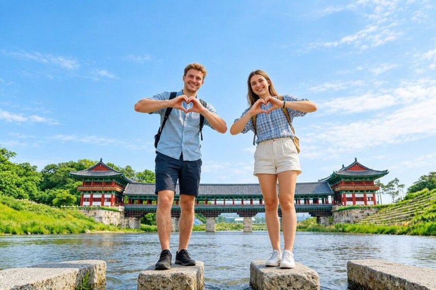 Two people making a heart shape with their hands in front of a traditional building by a water body. 1