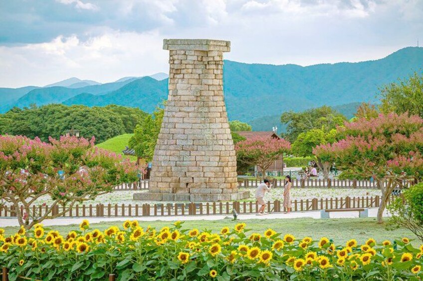 A stone tower, a wooden fence, and sunflowers in a park with mountains in the background. 3