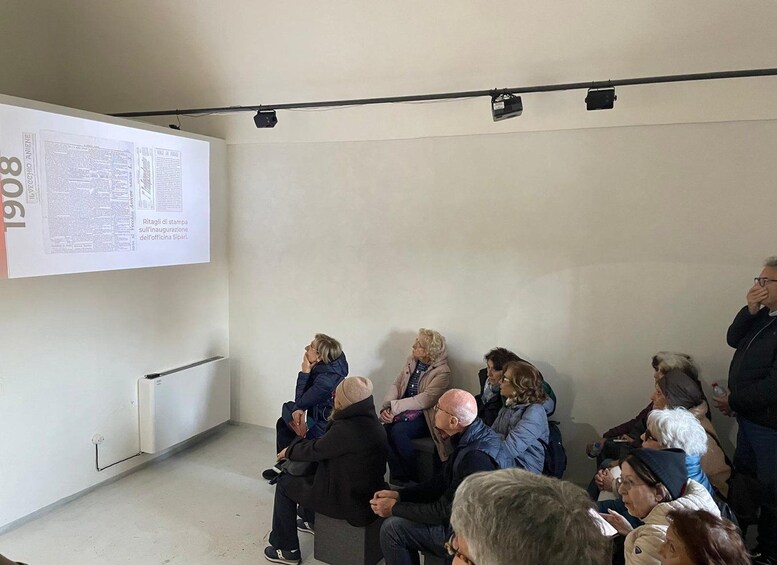 Un grupo de personas sentadas en una sala con una pantalla de proyección que muestra texto e imágenes. 1
