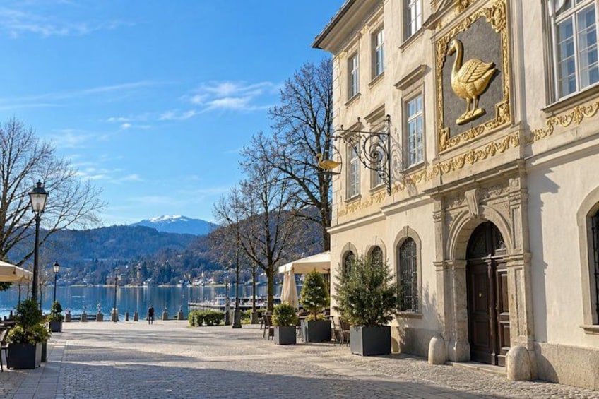 A cobblestone square with a building featuring a golden swan emblem, a lake, and mountains in the background. 3