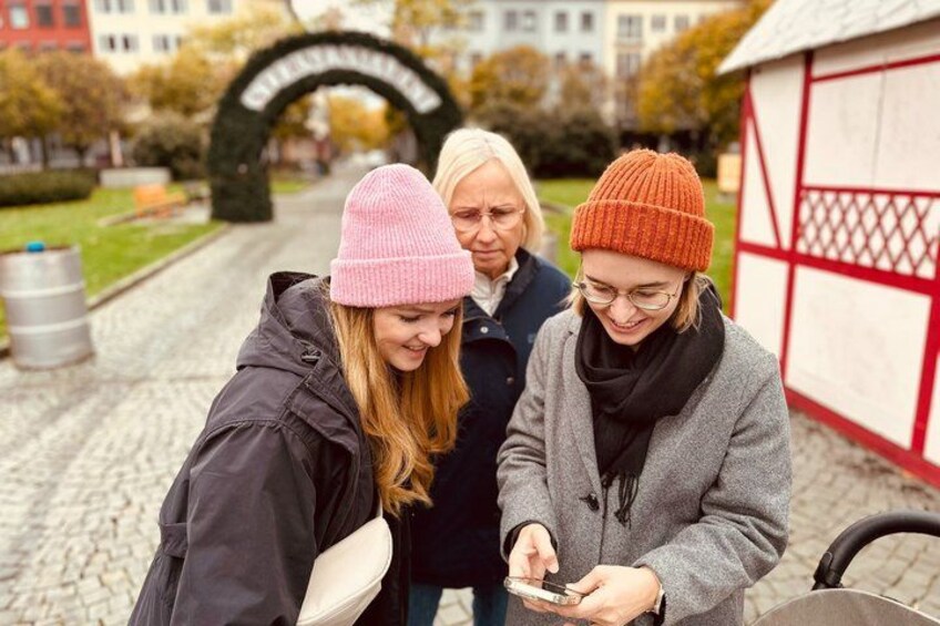 Three people standing outdoors near a building with a red and white facade, a cobblestone path, and a green park in the background. 1