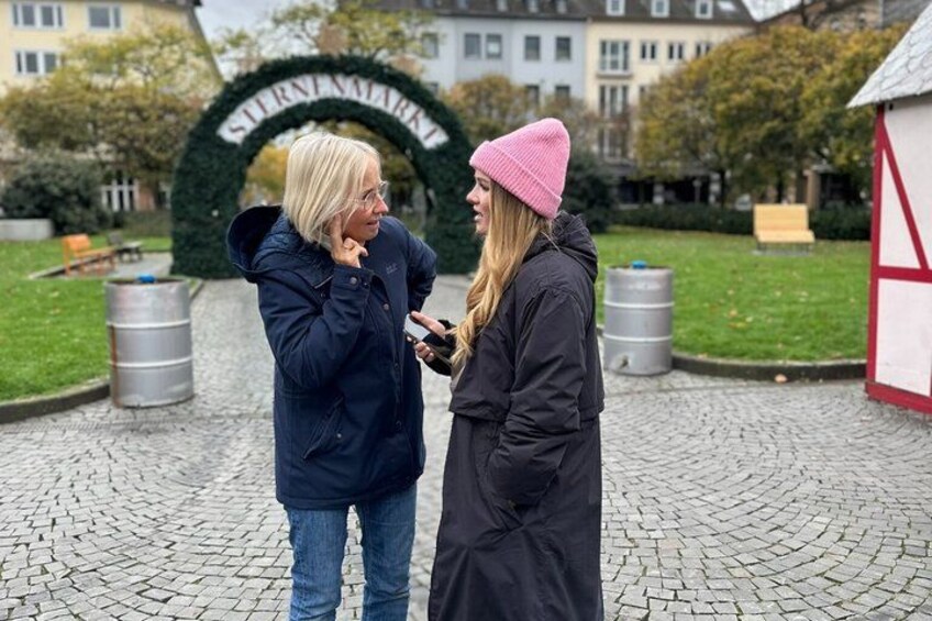 Two people conversing in a cobblestone square with a green archway and a building in the background. 5