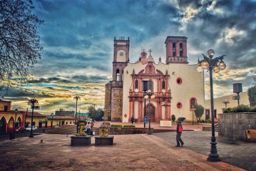 A historic church with a bell tower, a plaza with benches, and a cloudy sky. 1
