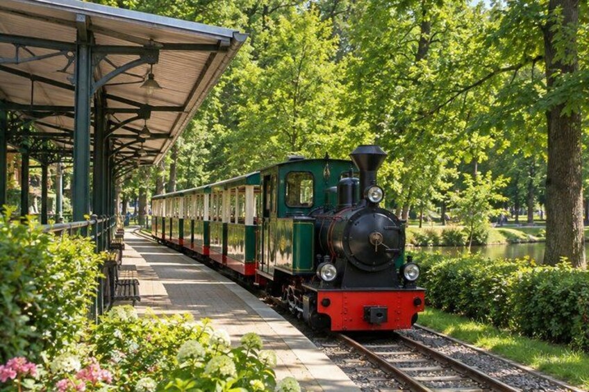 A green and red steam train at a station surrounded by lush greenery and a lake. 4