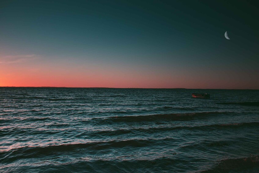 A calm sea at sunset with a boat in the distance and a crescent moon in the sky. 2