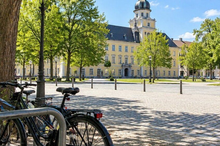 A paved square with bicycles parked, trees, and a historic building with a tower in the background. 2