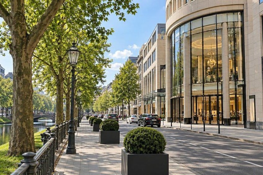 A tree-lined urban street with modern buildings, a canal, and parked cars. 3