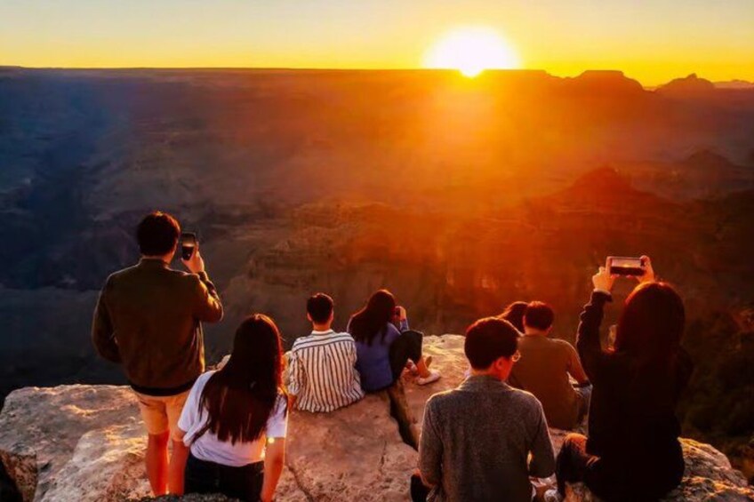 A group of people sitting on a rock overlooking a canyon with a sunset in the background. 1