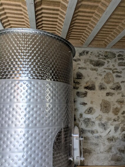 A metallic container with a textured surface and a valve, positioned against a stone wall with a wooden ceiling. 3