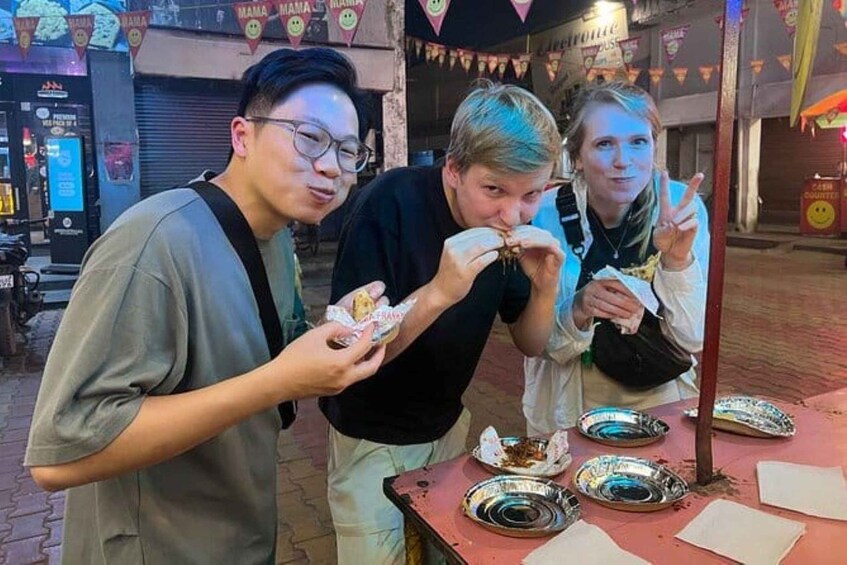 Trois personnes sont assises à une table rouge, devant un étal de nourriture en plein air, et mangent dans des assiettes en métal en utilisant des serviettes en papier. 5