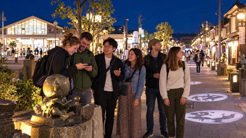 Un grupo de personas de pie en una calle de la ciudad de noche, con edificios iluminados y farolas al fondo. 2