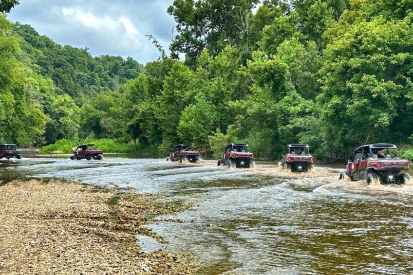 Four off-road vehicles crossing a shallow river in a forested area. 4