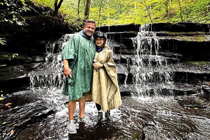A waterfall in a forested area with a person in a green shirt and another in a beige coat standing in the water. 3