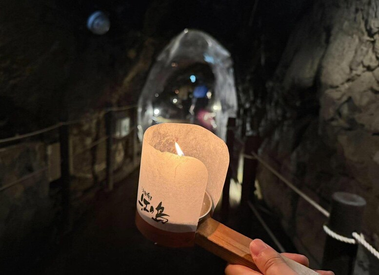 A hand holding a lit candle with Japanese characters, in a dimly lit cave with a rope barrier. 2