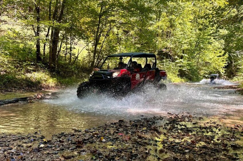 A red off-road vehicle crossing a shallow stream in a forested area. 5