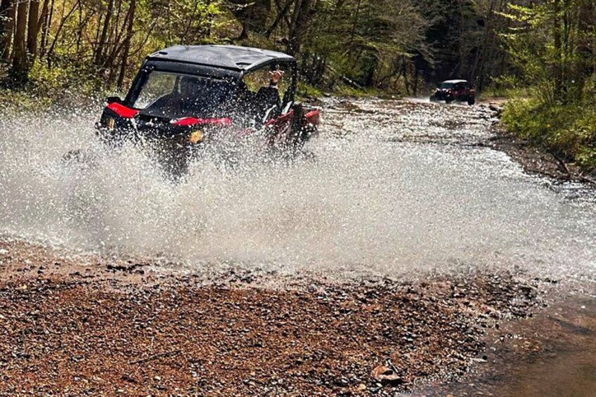 A vehicle driving through a shallow water crossing in a forested area. 3
