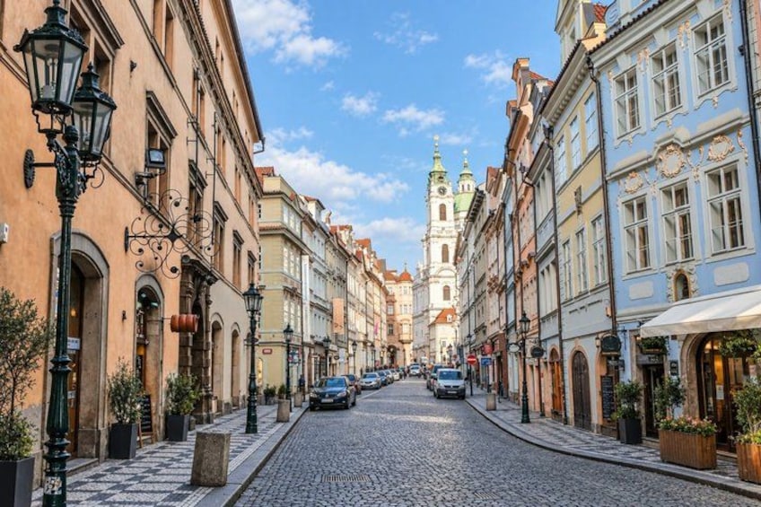 A cobblestone street lined with historic buildings, a church spire in the distance, and a clear blue sky. 5