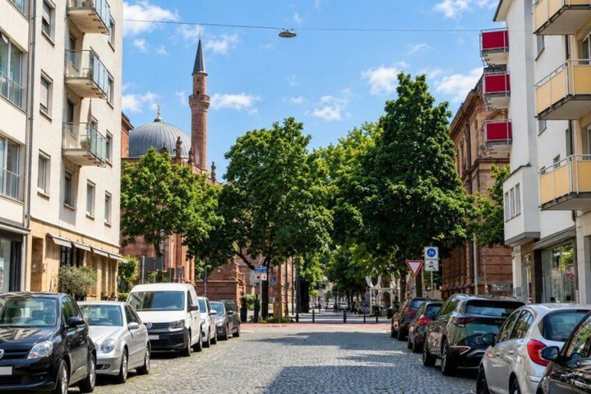 A cobblestone street lined with parked cars, buildings, and a mosque with a minaret in the background. 2