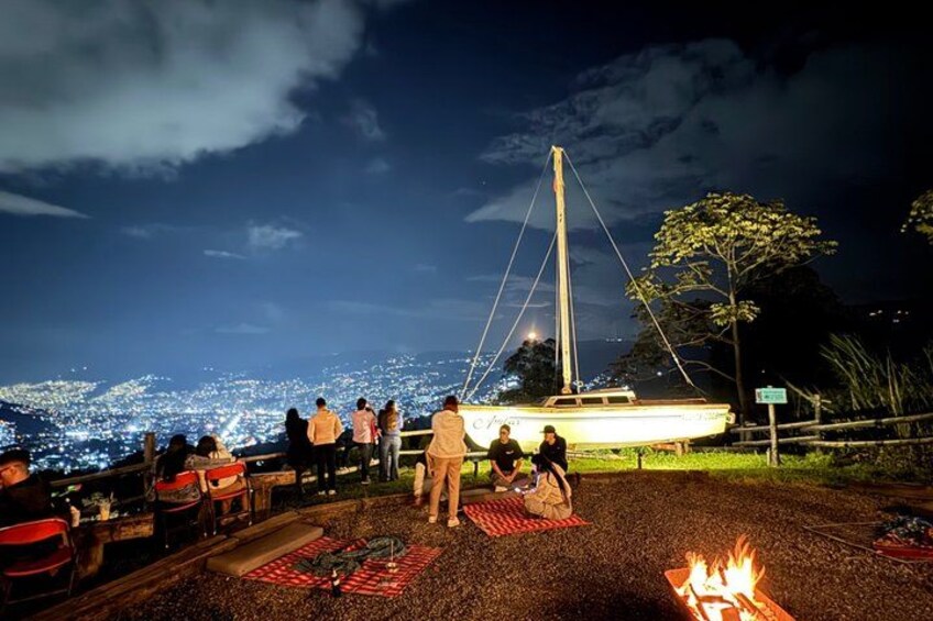 A group of people gathered around a campfire at night, overlooking a cityscape with a sailboat in the distance. 4