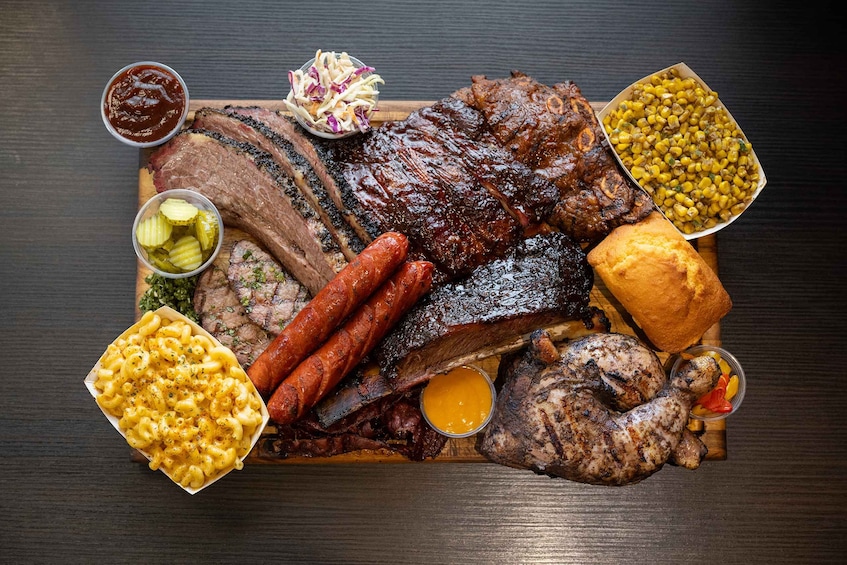 Variedad de carnes a la parrilla, incluyendo costillas, salchichas y un chuletón de cerdo, acompañadas de guarniciones como ensalada de col, macarrones con queso, maíz y pepinillos. 1