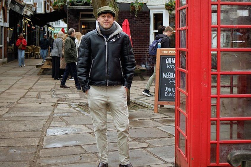 A person standing in front of a red telephone booth on a cobblestone street with shops and people in the background. 2