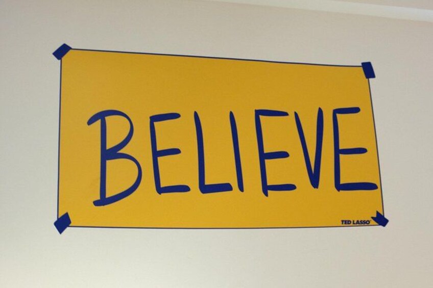 A yellow sign with the word 'BELIEVE' taped to a wall. 1