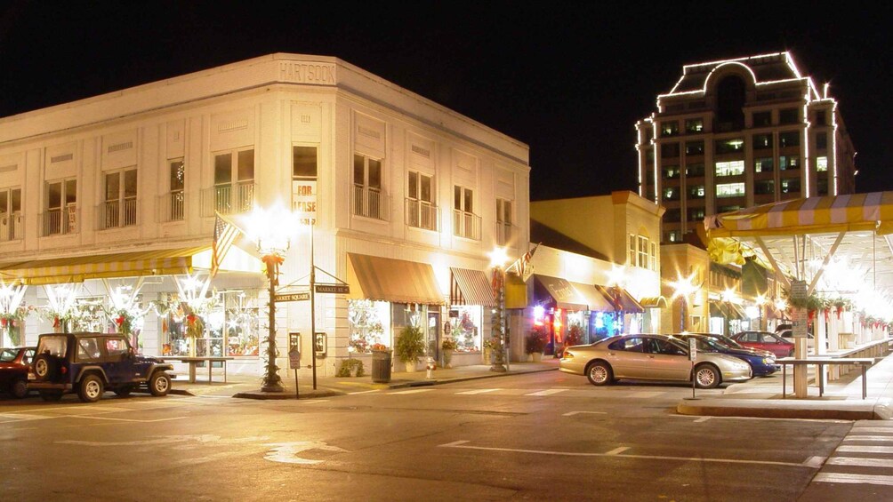A nighttime street scene with illuminated buildings, parked cars, and a pedestrian crossing. 2